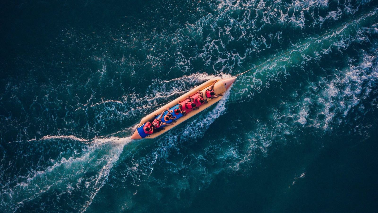 A bright banana boat full of riders in life jackets, seen from above on deep teal water